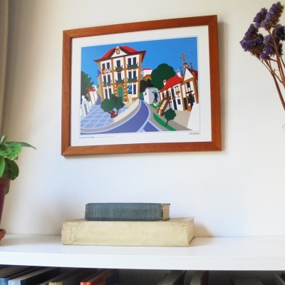 Quadro colorido emoldurado com ilustração de casas e árvores na parede sobre prateleira com livros e plantas.