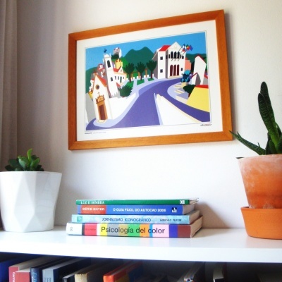 Quadro colorido de vila em moldura de madeira sobre parede branca, prateleira com livros e plantas em vasos