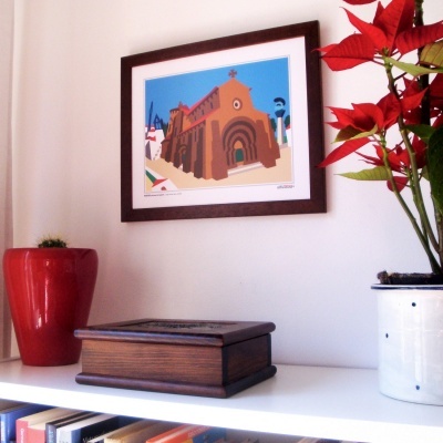 Quadro colorido de igreja em moldura de madeira, estante com caixa de madeira, vaso vermelho com planta, vaso branco com flores vermelhas