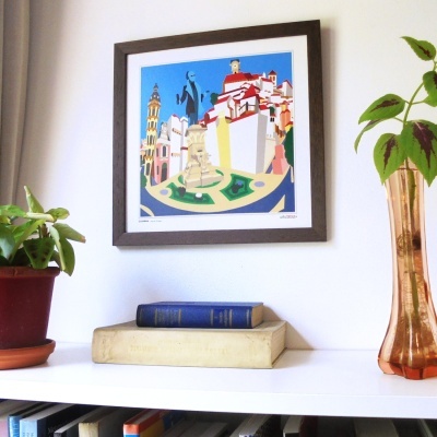 Quadro colorido com desenho da praça de Coimbra em moldura de madeira pendurado na parede, com plantas e livros numa prateleira branca abaixo.