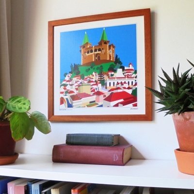 Quadro colorido com castelo e casas, moldura de madeira, sobre estante com plantas e livros.