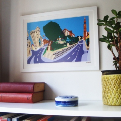 Quadro colorido em moldura branca sobre prateleira com livros, vaso amarelo e caixa redonda azul e branca