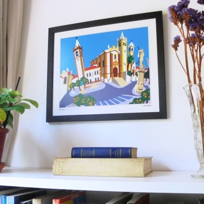 Quadro colorido de edifício histórico emoldurado na parede, sobre estante com livros e plantas