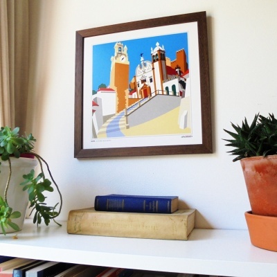 Quadro com ilustração colorida de edifícios pendurado numa parede branca sobre estante com livros e plantas.