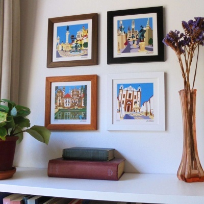 Quatro quadros de paisagens urbanas em molduras coloridas numa parede branca acima de estante com livros, planta e jarro com flores secas.