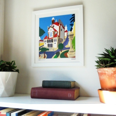 Quadro colorido de igreja com moldura branca acima de prateleira com livros e plantas