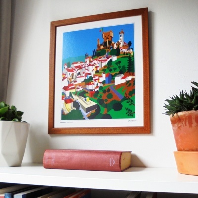 Quadro colorido com paisagem urbana em moldura de madeira, estante branca com livro e plantas