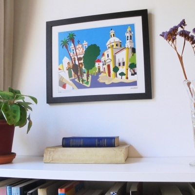 Quadro colorido de igreja com moldura preta na parede, livros e plantas numa prateleira branca