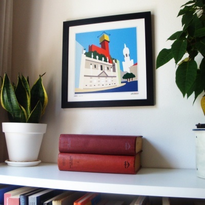 Quadro colorido de farol vermelho em moldura preta na parede, plantas e livros em estante branca