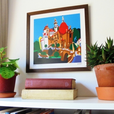 Gravura de edifício com torres coloridas em moldura de madeira, plantas em vasos de terracota e livros numa prateleira branca.