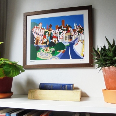 Quadro colorido com moldura de madeira escura pendurado na parede, plantas em vasos e livros em estante branca.