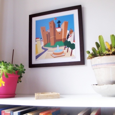 Quadro com ilustração de castelo pendurado na parede, estante branca com vasos de plantas e cactos