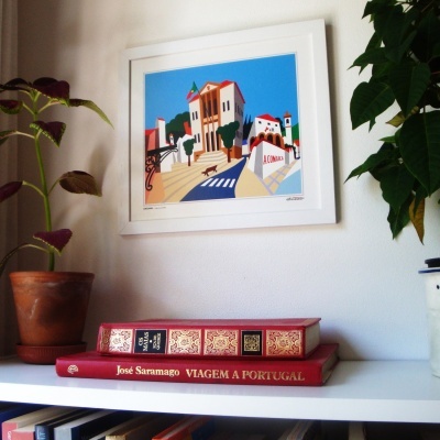 Impressão colorida emoldurada com casa urbana portuguesa, plantas em vaso e livros de José Saramago numa prateleira branca