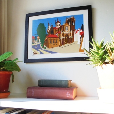 Quadro colorido de edifícios em moldura preta sobre prateleira com livros e plantas