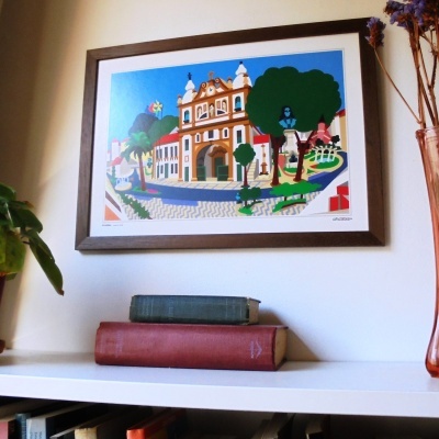 Gravura colorida de edifício histórico com árvores em moldura castanha na parede acima de estante branca com dois livros.