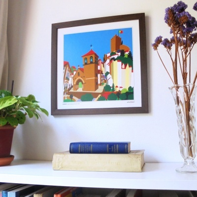 Quadro colorido de edifícios com céu azul numa parede branca sobre estante com livros e plantas