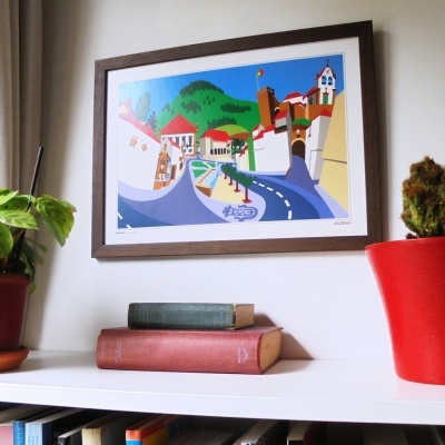 Quadro colorido de vila com estrada numa parede branca, plantas em vasos e livros numa estante.