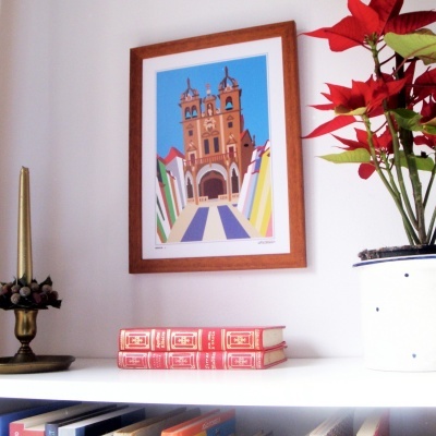 Estante branca com livros, vela dourada, planta com folhas vermelhas e quadro com ilustração de igreja.
