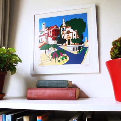 Quadro colorido de cidade estilizada em moldura branca acima de prateleira com livros e vasos de plantas