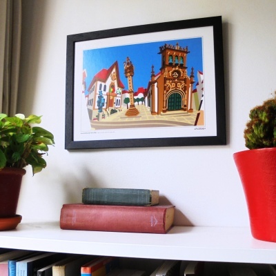 Quadro colorido de edifícios históricos com moldura preta, plantas em vasos vermelhos, livros antigos numa prateleira branca.