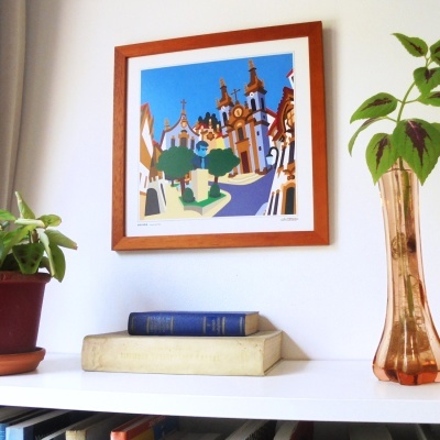 Quadro de madeira com ilustração colorida de uma praça histórica pendurado numa parede branca ao lado de plantas e livros