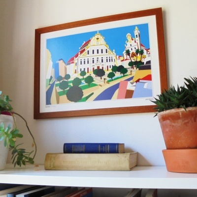 Moldura com ilustração colorida de edifícios e árvores pendurada em parede branca, com livros e vasos de plantas numa prateleira abaixo.