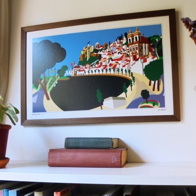 Quadro colorido de Alcácer do Sal em moldura madeira, sobre estante branca com livros antigos e plantas