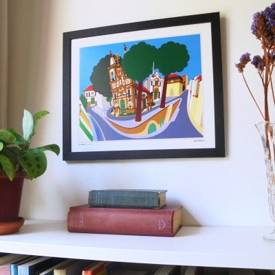 Quadro colorido emoldurado com cena urbana estilizada, prateleira branca com livros e flores secas.