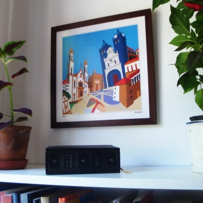 Quadro colorido de edifícios numa parede com plantas e livro em prateleira