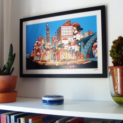 Quadro a cores de cidade pendurado numa parede acima de prateleira com plantas e recipiente azul e branco