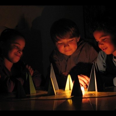 quatro crianças sentadas em torno de prismas triangulares iluminados numa mesa