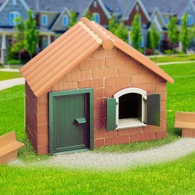 Casa para cães plástica estilizada como casa de tijolos com porta e janelas verdes, com bancos de madeira e relvado