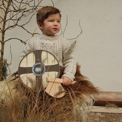 Criança com suéter de malha bege segurando escudo de madeira redondo, sentada num local com plantas secas e parede branca.