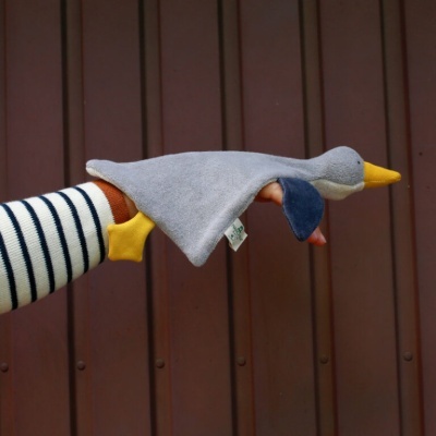 Fantoche de mão em forma de pato cinzento com bico e patas amarelas, orelhas azuis, em fundo de porta de madeira castanha