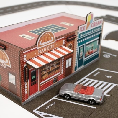 Modelo de loja em papel com fachada vermelha, toldo às riscas brancas e vermelhas, placa BAKERY e carrinho prata no estacionamento.