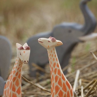 Duas girafas de madeira com padrão laranja e branco e elefantes cinzentos ao fundo sobre palha seca