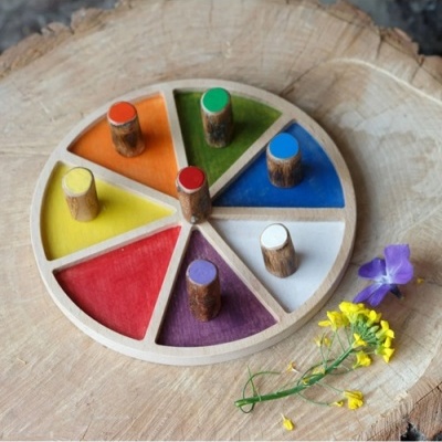 Jogo educativo redondo de madeira com segmentos coloridos e peças cilíndricas, sobre mesa rústica com flores.