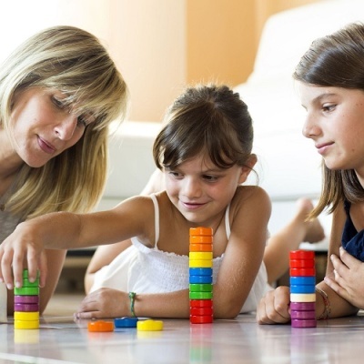 Jogo de empilhar peças coloridas com duas crianças e uma mulher sentadas no chão.