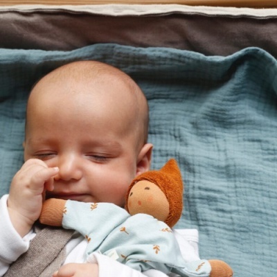 Bebé a dormir a abraçar uma boneca de tecido com roupa azul clara e gorro laranja sobre um pano azul turquesa.