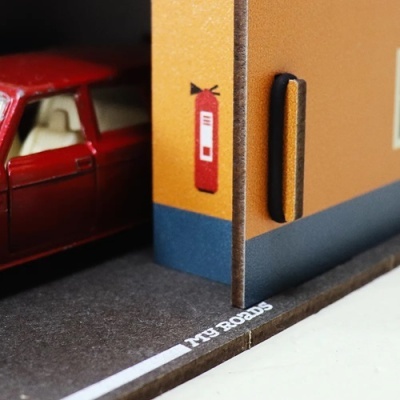 Maqueta de rua com parede laranja, extintor vermelho pintado, porta preta, texto TYB BOARDS no chão e carro vermelho ao fundo