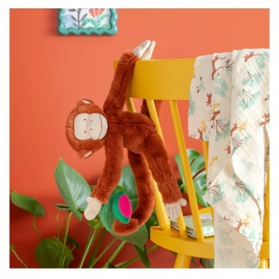 Pelúcia de macaco castanho pendurado numa cadeira amarela com pano decorado e plantas ao fundo