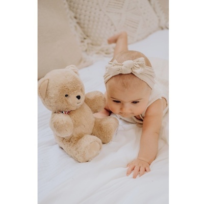 Bebé com faixa creme e vestido branco a olhar para ursinho de peluche castanho numa cama branca