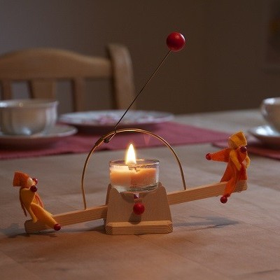 Castiçal em forma de baloiço com vela acesa e figuras decorativas em madeira numa mesa de jantar.