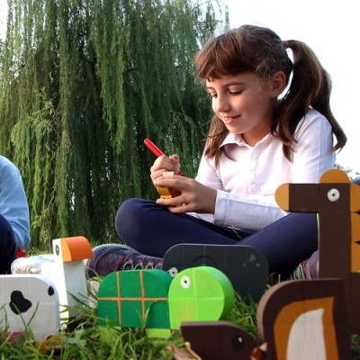 Criança a pintar animais de madeira coloridos sentada na relva com árvore ao fundo