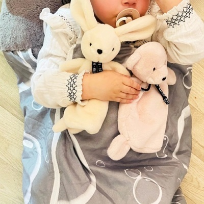 Bebé com bata branca segurando peluches de coelho e porco sobre cobertor cinzento com padrão branco