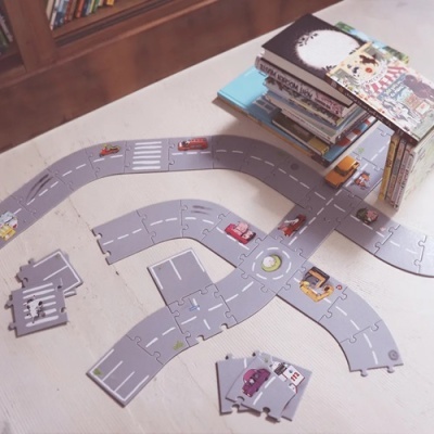 Peças de estrada de brinquedo cinza e carros coloridos numa superfície clara com livros ao fundo