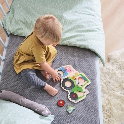 Criança a brincar com puzzle de madeira colorido numa cama com colcha cinzenta e almofada verde clara