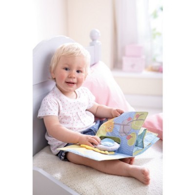 Criança sentada numa cama branca, a ler livro de tecido colorido.