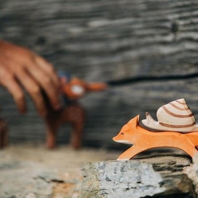 Figura de raposa em madeira pintada laranja com padrão branco, sobre pedra, com mão ao fundo segurando outra figura
