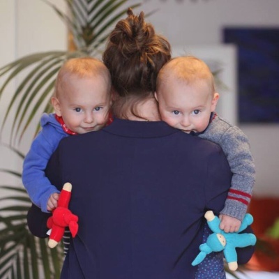 Mulher de costas a segurar dois bebés, um com boneco de peluche vermelho e outro com azul.